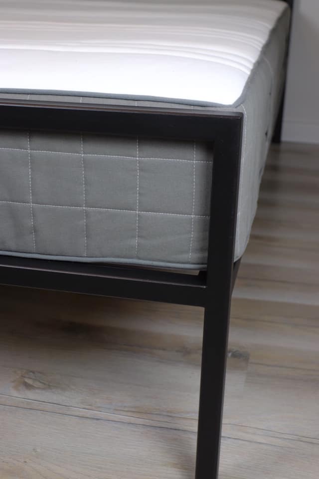 Szczegół minimalistycznego łóżka z szarą, pikowaną ramą i białym materacem, na drewnianej podłodze.