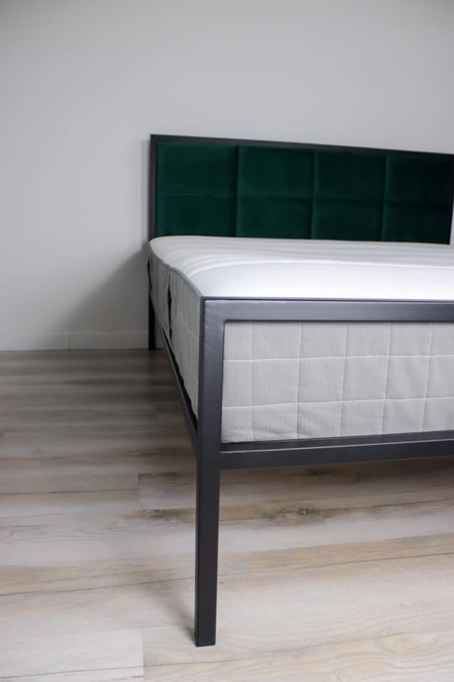 Łóżko z czarną ramą i zielonym, tapicerowanym wezgłowiem w stylu Chesterfield, ustawione na panelach podłogowych imitujących drewno.