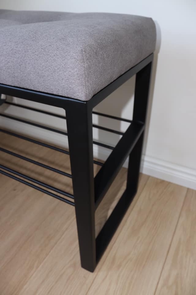 Tapicerowana ławka z szarą poduszką i czarną metalową ramą, ustawiona na drewnianej podłodze przy białej ścianie z listwą przypodłogową.