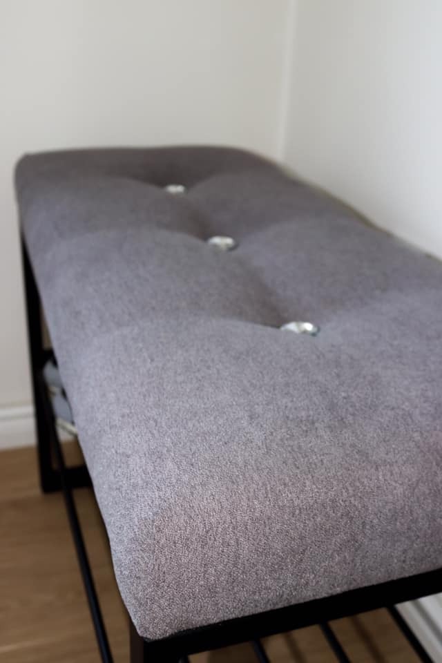 Szary tapicerowany puf z trzema srebrnymi guzikami na metalowej ramie z półką na buty, ustawiony w rogu pomieszczenia.