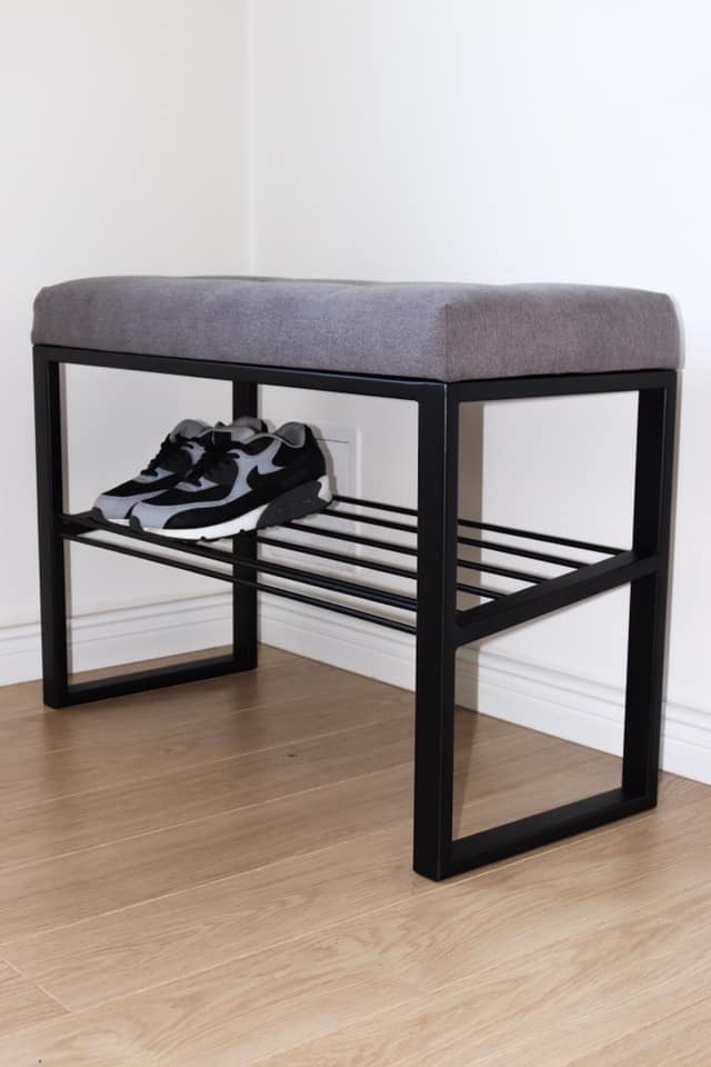 Ławka do przedpokoju z szarą tapicerowaną poduszką siedziska i czarną metalową ramą z półką na buty, na której leżą czarno-szare sportowe buty.