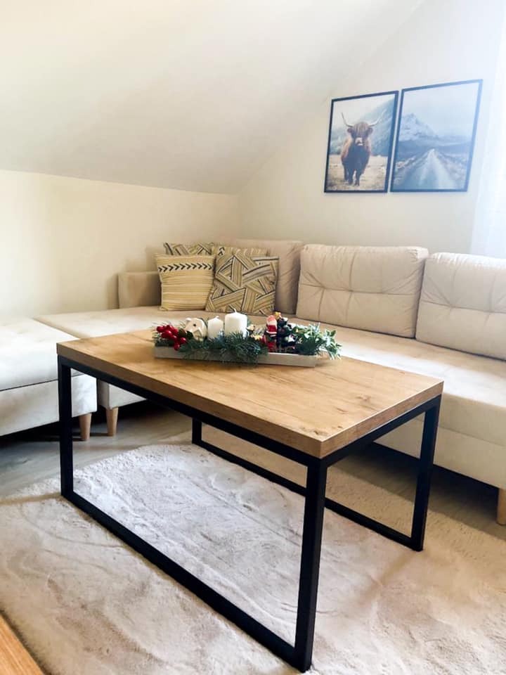 Stolik kawowy z blatem w odcieniu dębu klasycznego i czarnymi, metalowymi nogami, ustawiony na puszystym, białym dywanie w salonie z beżową sofą narożną i dekoracjami świątecznymi na blacie.