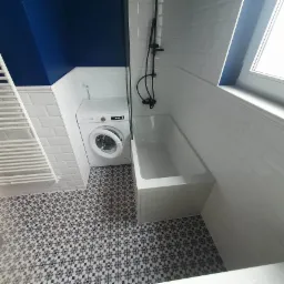 Nowoczesna łazienka z białą wanną, pralką z otwartymi drzwiczkami, grzejnikiem drabinkowym i wzorzystą podłogą w odcieniach szarości, wykończona białymi płytkami typu metro i ścianą pomalowaną...