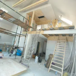 Poddasze w trakcie adaptacji, z widoczną konstrukcją drewnianą, rusztowaniami, płytami karton-gips i dwiema drabinami, oraz fragmentem ocieplenia podłogi i rozrzuconymi materiałami budowlanymi.