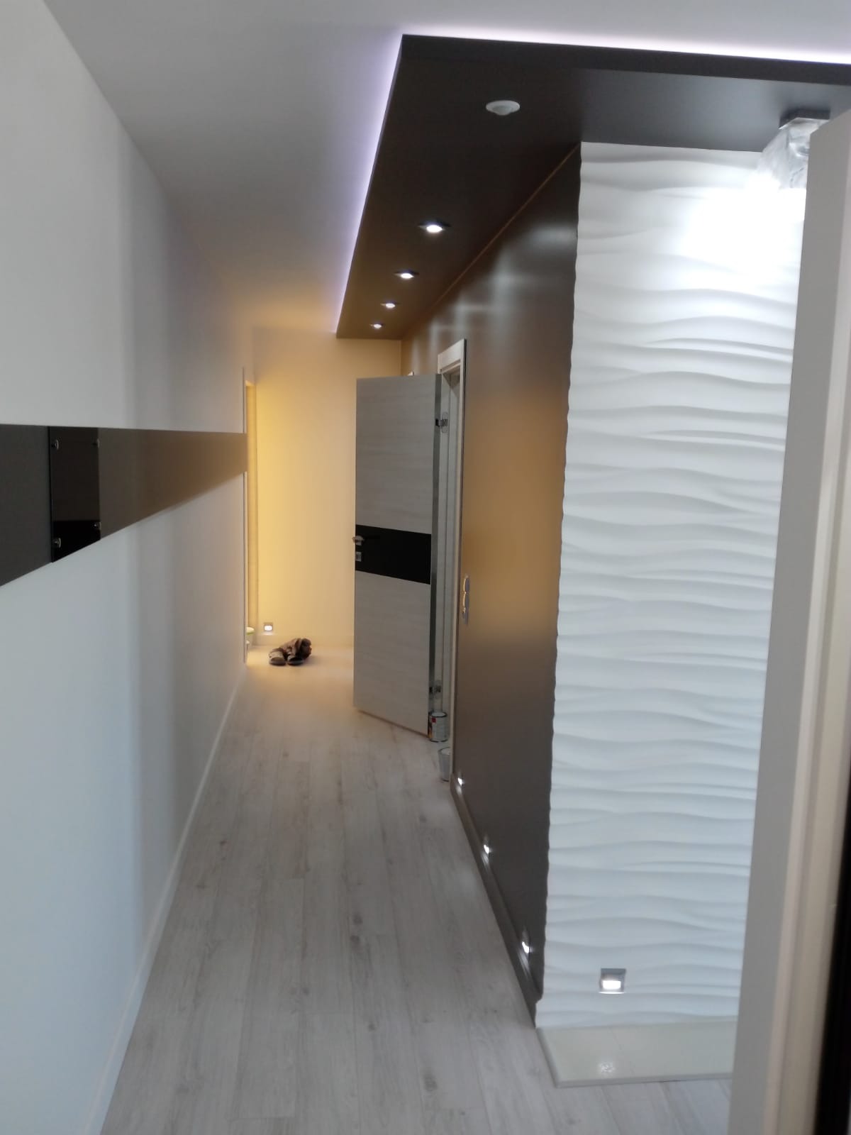 Korytarz po remoncie z oświetleniem LED w suficie i podłodze, dekoracyjna ściana z białymi panelami 3D, otwarte drzwi z czarnym akcentem, widoczne buty na podłodze.