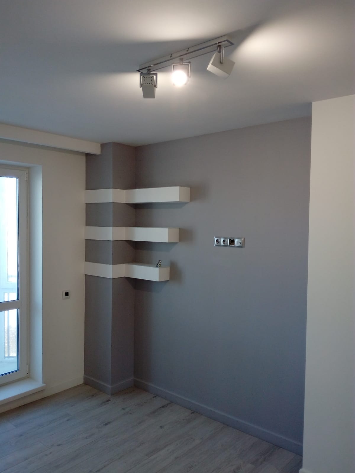 Wykończone wnętrze w Warszawie: szara ściana z trzema białymi, narożnymi półkami, szara podłoga imitująca drewno, białe okno z widokiem na zewnątrz oraz nowoczesne oświetlenie sufitowe.