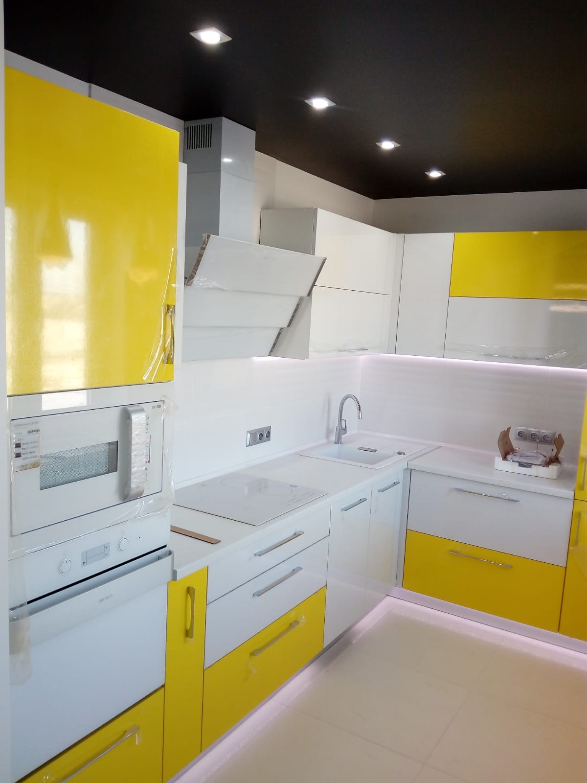 Nowoczesna kuchnia w bieli i żółci z błyszczącymi frontami, jeszcze w folii ochronnej, widoczny sprzęt AGD Gorenje i czarny sufit z punktowym oświetleniem.