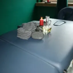 Przygotowany stół do masażu z kamieniami, stosem szarych ręczników, olejkiem w szklanym naczyniu i zapaloną świecą, na tle zielonej ściany.