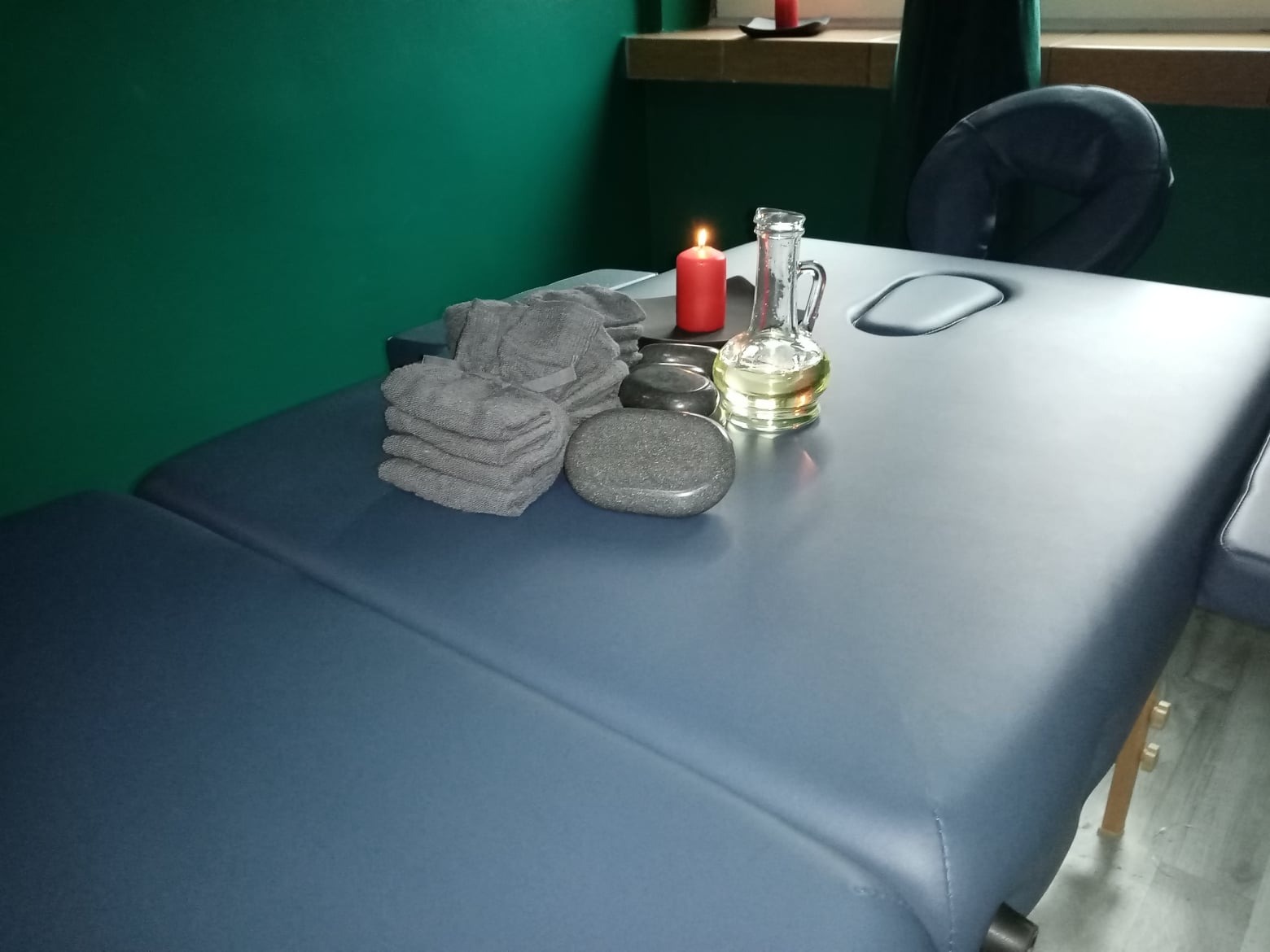 Przygotowany stół do masażu z kamieniami, stosem szarych ręczników, olejkiem w szklanym naczyniu i zapaloną świecą, na tle zielonej ściany.