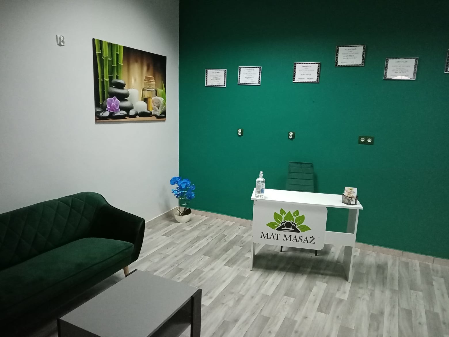 Recepcja w gabinecie masażu z zieloną ścianą, dyplomami, obrazem z motywem zen, kanapą i stolikiem kawowym.