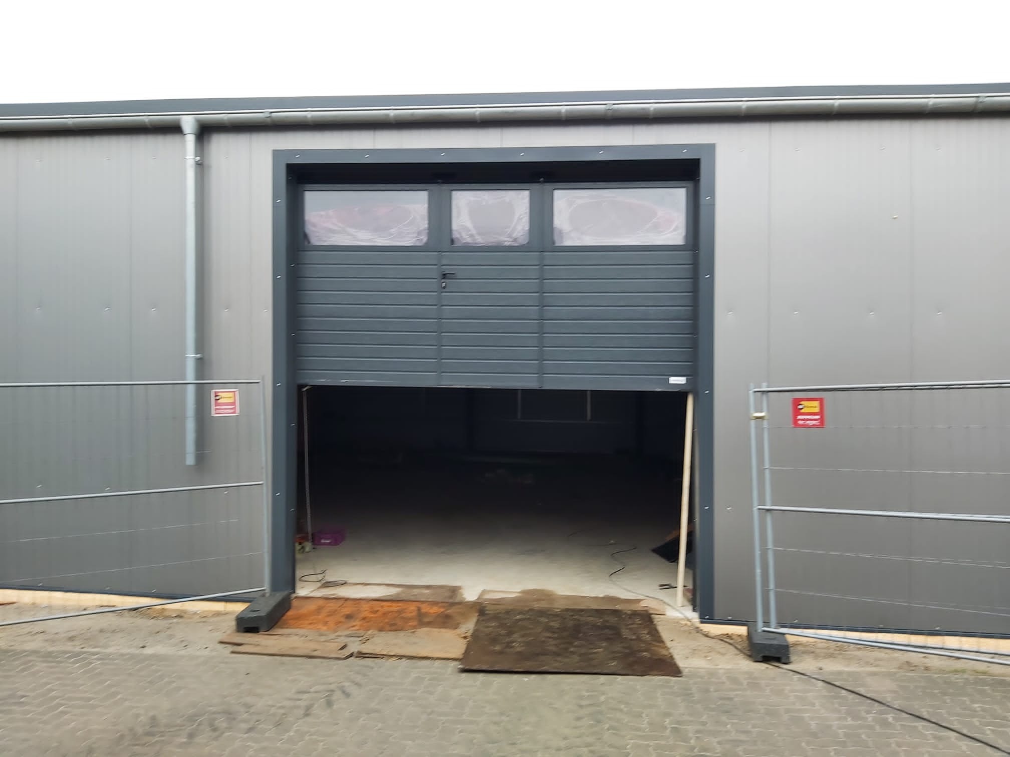 Szara hala przemysłowa z częściowo otwartą segmentową bramą garażową z trzema oknami w górnej części, zabezpieczona barierkami ostrzegawczymi.