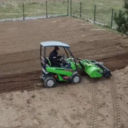 Zielony traktor z glebogryzarką wyrównuje teren pod przyszły trawnik, widok z góry, w tle ogrodzenie posesji.