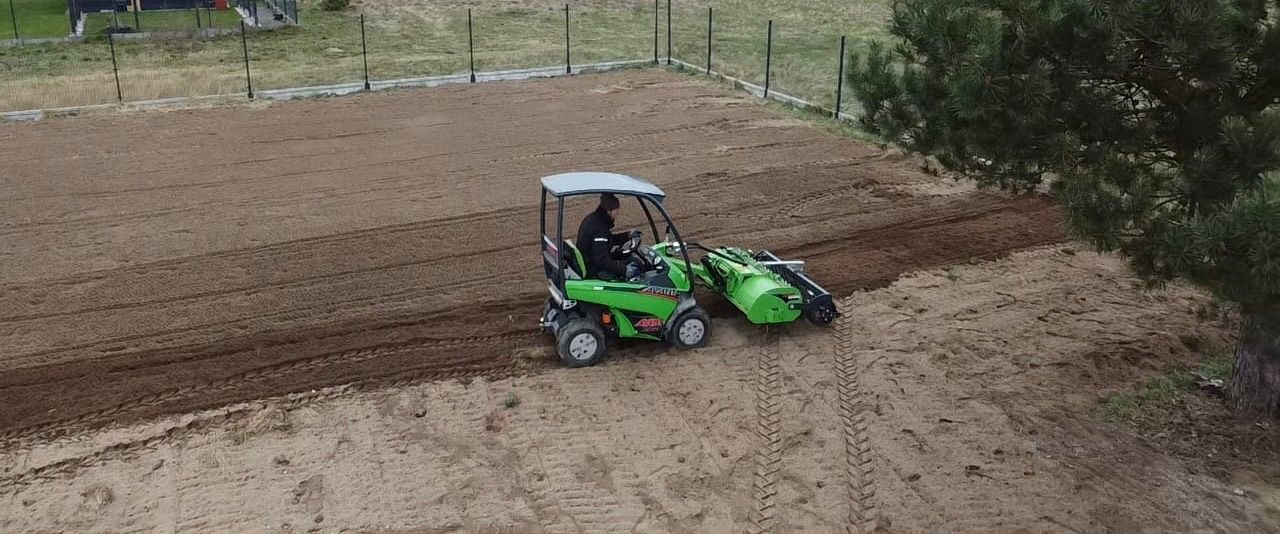Zielony traktor z glebogryzarką wyrównuje teren pod przyszły trawnik, widok z góry, w tle ogrodzenie posesji.
