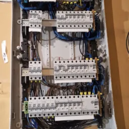 Otwarta skrzynka bezpiecznikowa z widocznymi bezpiecznikami, przewodami w kolorach niebieskim, czarnym i brązowym oraz listwą zaciskową w tle, zamontowana na ścianie z widocznymi śladami po...