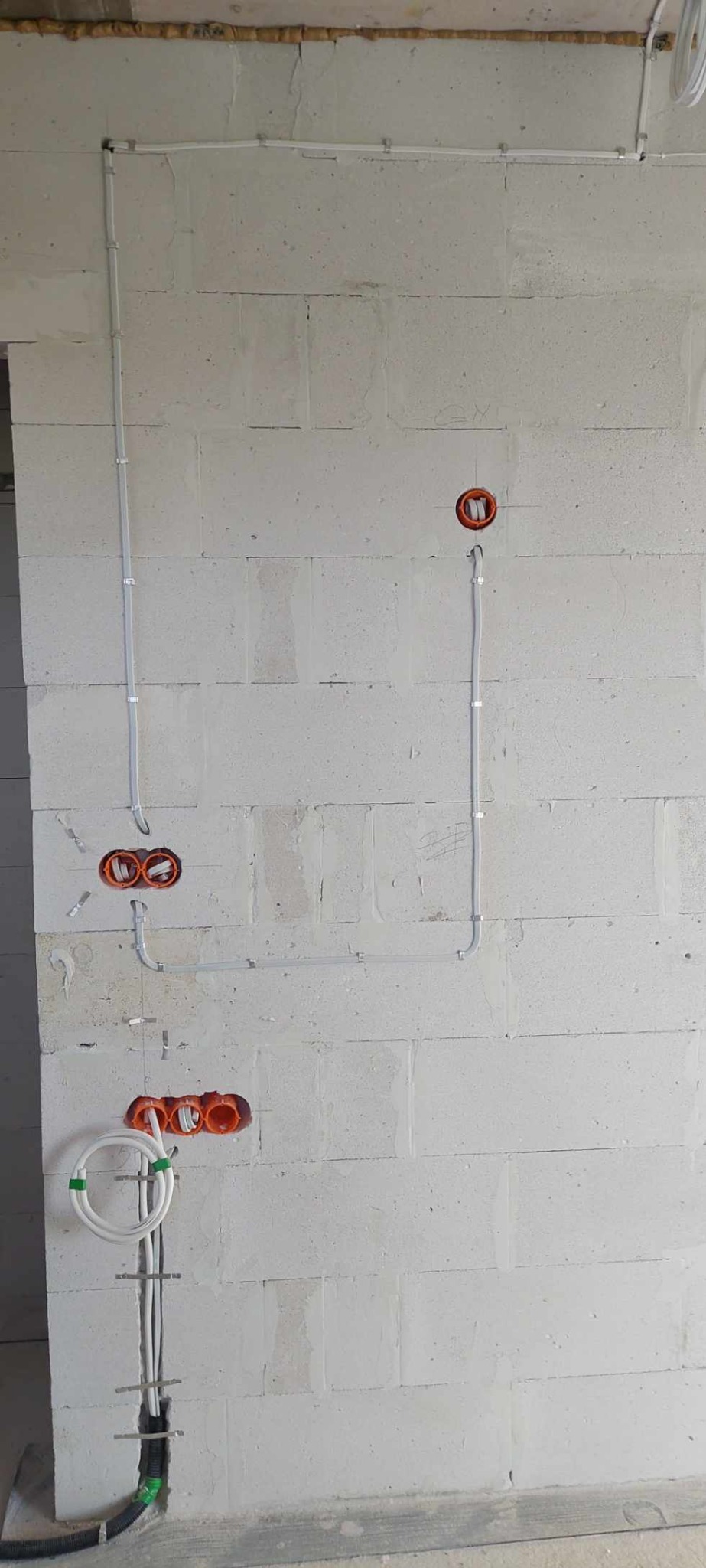 Surowa ściana z betonu komórkowego z zamontowanymi puszkami elektrycznymi i poprowadzonymi przewodami w białych listwach mocowanych klamrami.