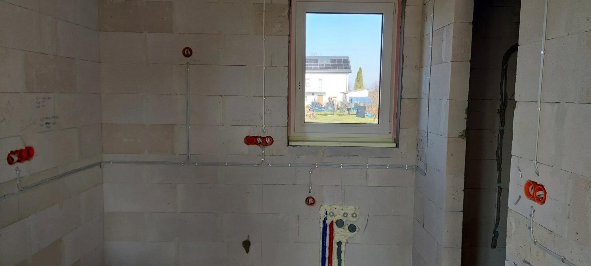 Świeżo położone przewody elektryczne w surowym wnętrzu, widoczne puszki instalacyjne w ścianach z betonu komórkowego, okno z widokiem na dom z panelami słonecznymi.