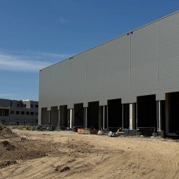 TEMAX Sp. Z O.O. - Wznoszony budynek magazynowy z widoczną elewacją z paneli i otworami na bramy, teren budowy z hałdą piasku w pierwszym planie, błękitne niebo w tle.