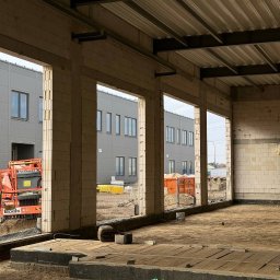 TEMAX Sp. Z O.O. - Wnętrze hali w trakcie budowy, widoczne surowe ściany z betonu komórkowego, stalowa konstrukcja dachu, podłoże w piasku i elementy izolacji, w tle budynek przemysłowy i podnośnik koszowy.