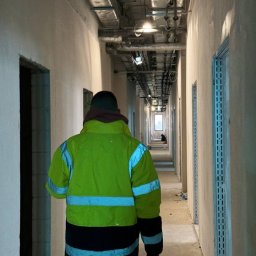 TEMAX Sp. Z O.O. - Pracownik w odblaskowej kurtce przechodzi korytarzem w trakcie remontu, widoczne surowe ściany przygotowane do wykończenia, instalacje pod sufitem i metalowe profile ościeżnic.