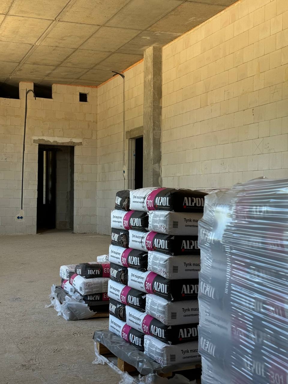 Stos worków z zaprawą tynkarską Alpol 301 na palecie w surowym wnętrzu budynku z pustaków betonowych, widoczne otwory drzwiowe i instalacja elektryczna.