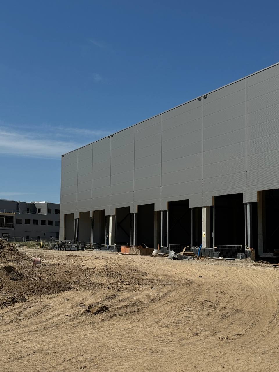 Wznoszony budynek magazynowy z widoczną elewacją z paneli i otworami na bramy, teren budowy z hałdą piasku w pierwszym planie, błękitne niebo w tle.