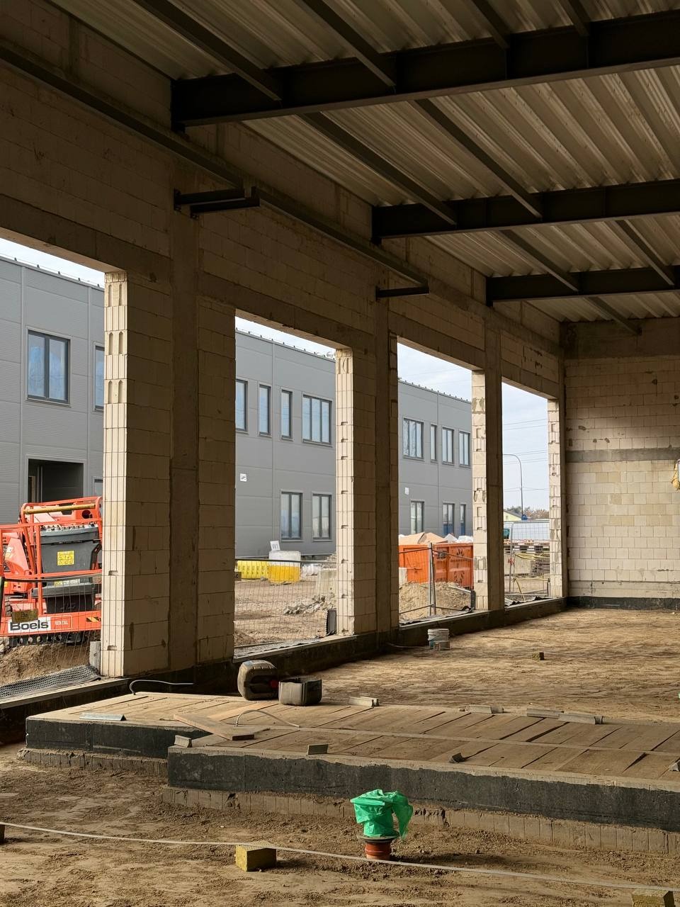 Wnętrze hali w trakcie budowy, widoczne surowe ściany z betonu komórkowego, stalowa konstrukcja dachu, podłoże w piasku i elementy izolacji, w tle budynek przemysłowy i podnośnik koszowy.