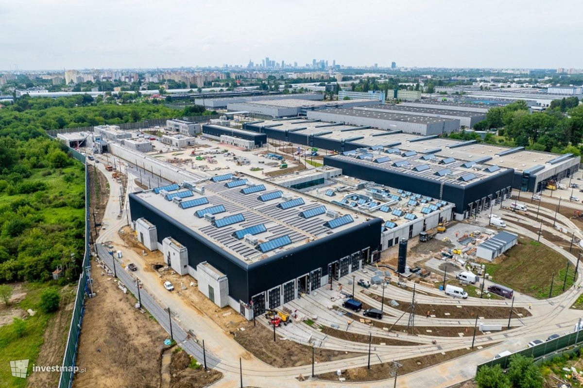 Panorama z lotu ptaka na plac budowy dużego kompleksu przemysłowego z częściowo ukończonymi budynkami, widoczne instalacje na dachach, teren przygotowany pod infrastrukturę, w tle zabudowa miejska...