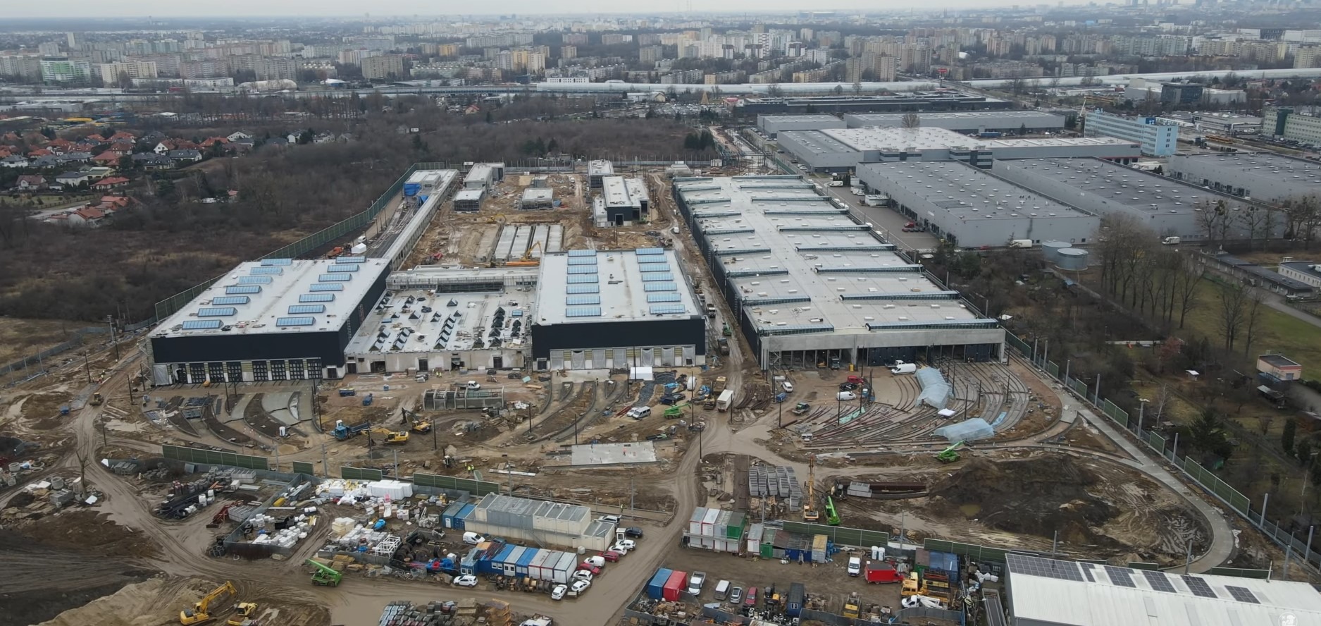 Rozległy widok z lotu ptaka na plac budowy w Warszawie, ukazujący budynki magazynowe w trakcie wznoszenia, z widocznymi elementami konstrukcyjnymi, sprzętem budowlanym i przygotowanym terenem.