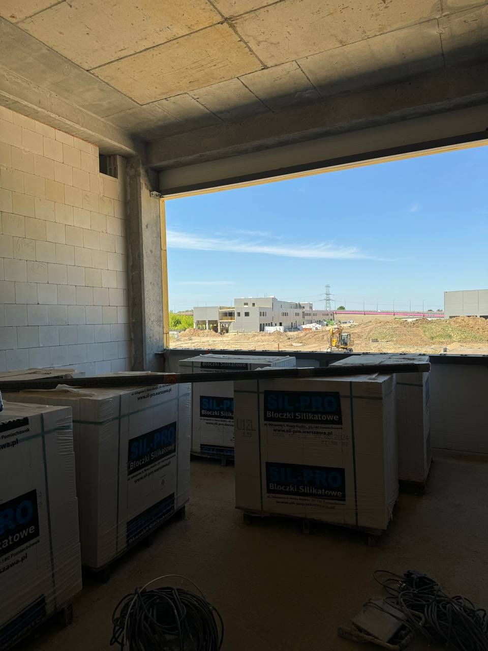 Wnętrze surowego budynku w trakcie budowy, widoczne palety z bloczkami silikatowymi Sil-Pro, ściana z białych bloczków, surowy betonowy strop i widok na plac budowy z żółtą koparką w tle przez...