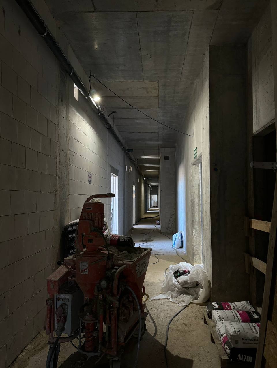 Wnętrze korytarza w stanie surowym z widoczną maszyną do tynkowania oraz workami z zaprawą tynkarską, ściany z bloczków, surowy betonowy strop, oświetlenie budowlane