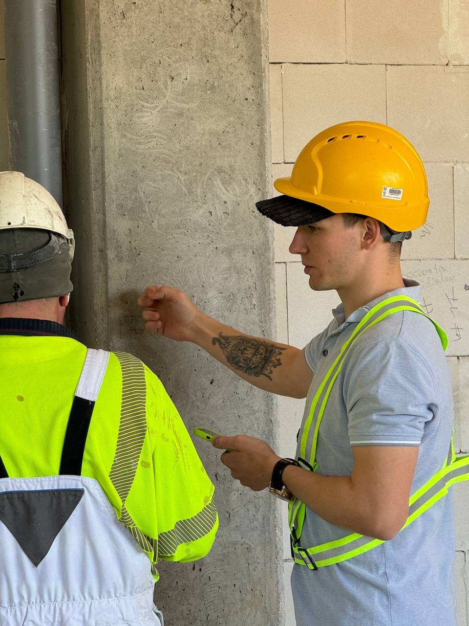 Dwóch mężczyzn w odblaskowych kamizelkach i kaskach ocenia jakość murowanej ściany z betonu komórkowego. Jeden z nich dotyka ściany, drugi trzyma telefon.