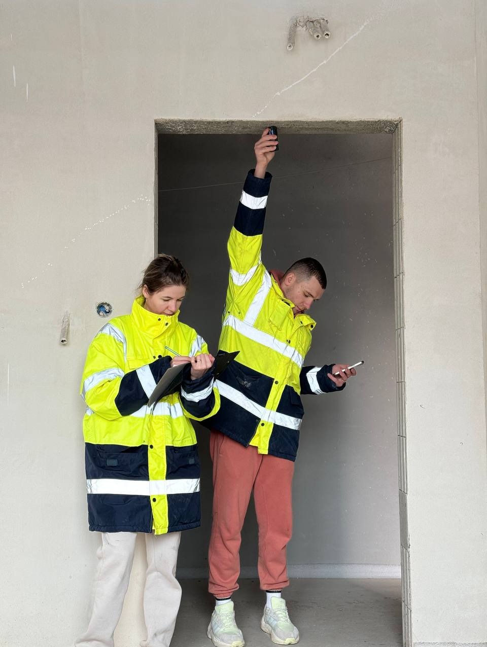 Dwoje pracowników w odblaskowych kurtkach, kobieta notuje na podkładce, mężczyzna mierzy wysokość otworu drzwiowego smartfonem, surowy stan ścian wewnątrz budynku.