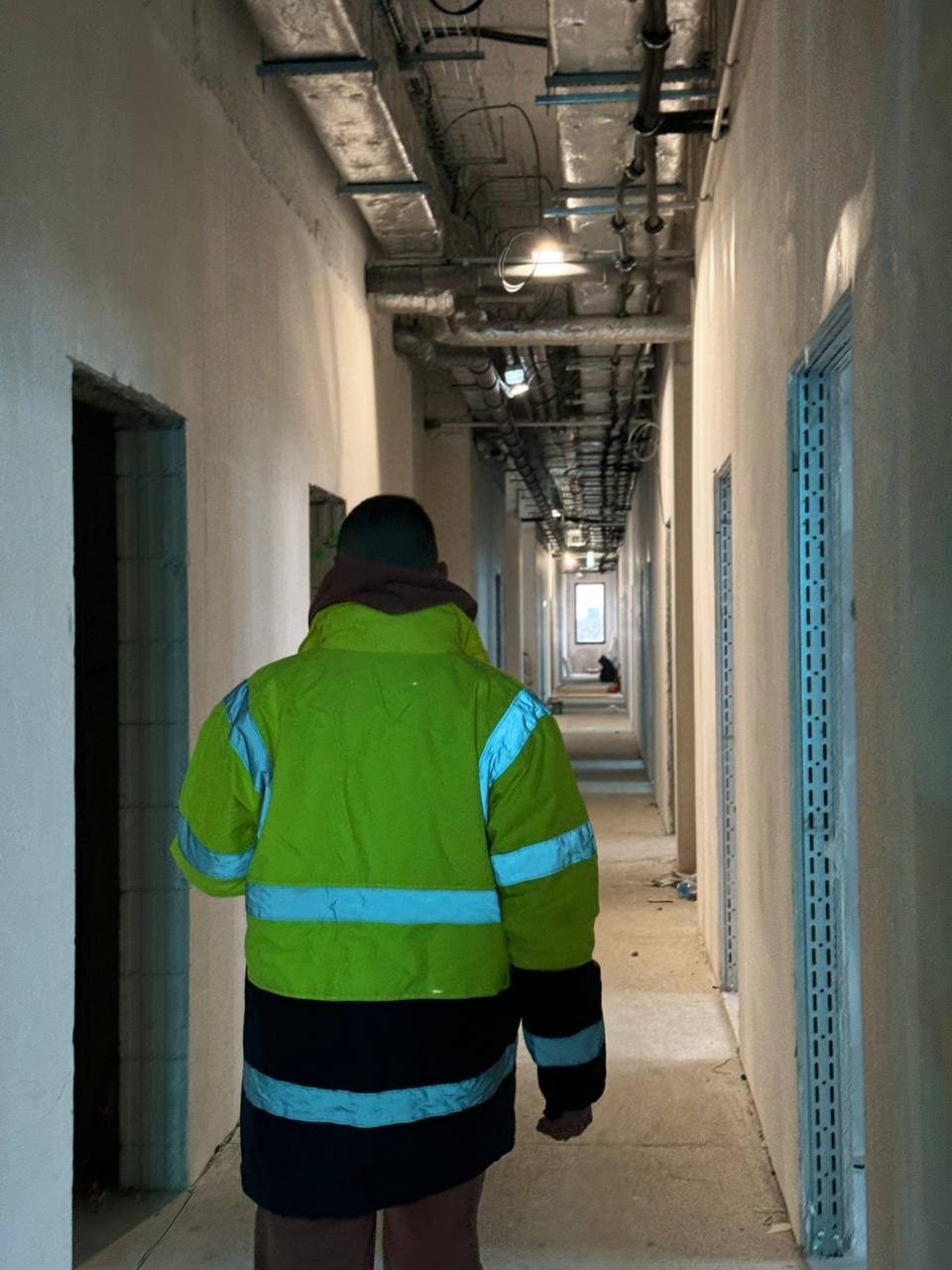 Pracownik w odblaskowej kurtce przechodzi korytarzem w trakcie remontu, widoczne surowe ściany przygotowane do wykończenia, instalacje pod sufitem i metalowe profile ościeżnic.