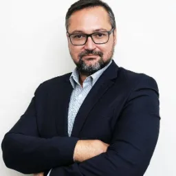 Portret mężczyzny w okularach, z brodą, ubranego w granatową marynarkę i błękitną koszulę, pozującego z założonymi rękami na jasnym tle.