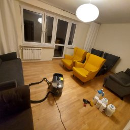 czyszczenie-kanap.pl - Wnętrze pokoju z ciemnoszarą sofą, dwoma żółtymi fotelami i sprzętem do czyszczenia tapicerki na drewnianej podłodze, widoczne okna z białymi ramami i jasne zasłony.