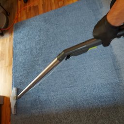 czyszczenie-kanap.pl - Czyszczenie niebieskiego dywanu za pomocą odkurzacza piorącego z metalową rurą i przezroczystą dyszą, widoczne drewniane panele podłogowe i fragment sofy.