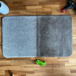 Dywanik z widoczną różnicą czystości: lewa strona jaśniejsza, prawa strona ciemniejsza, obok butelka z rozpylaczem, wiertarka z nakładką czyszczącą i zielony skrobak na drewnianej podłodze.