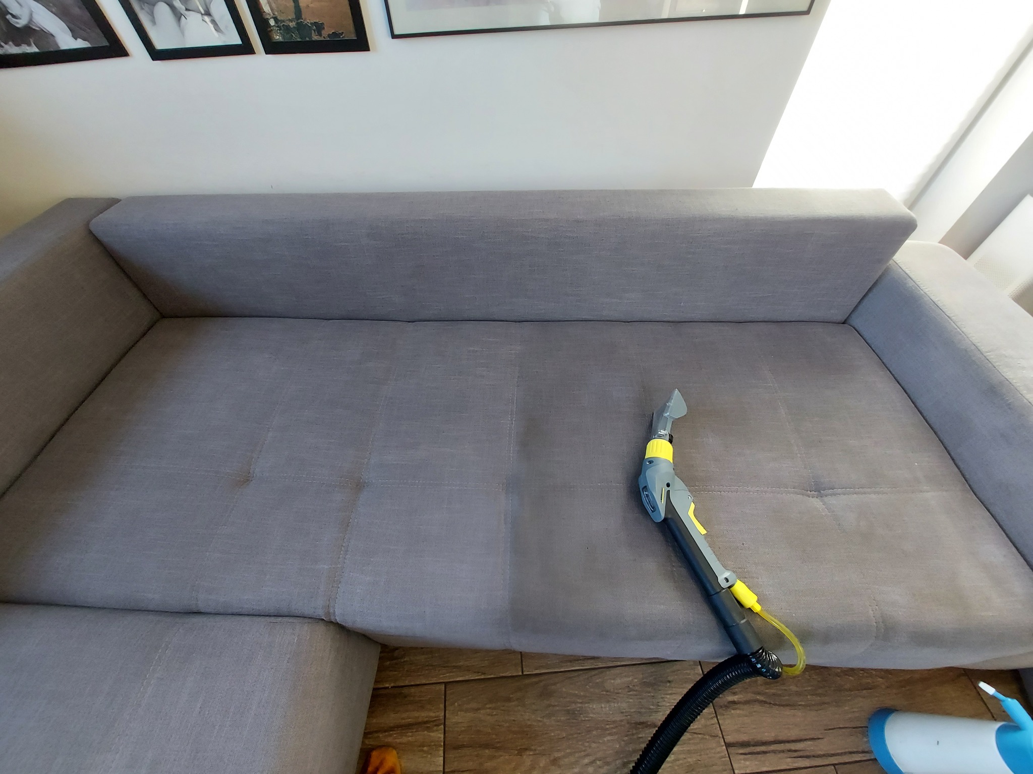 Szara sofa modułowa częściowo wyczyszczona, widoczna różnica w odcieniu, obok dysza parowa z odkurzaczem.