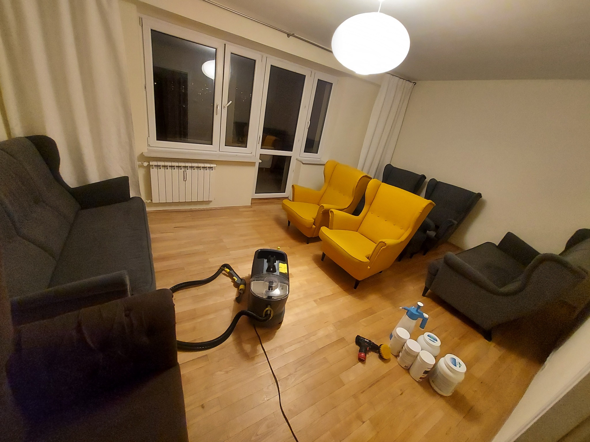Wnętrze pokoju z ciemnoszarą sofą, dwoma żółtymi fotelami i sprzętem do czyszczenia tapicerki na drewnianej podłodze, widoczne okna z białymi ramami i jasne zasłony.