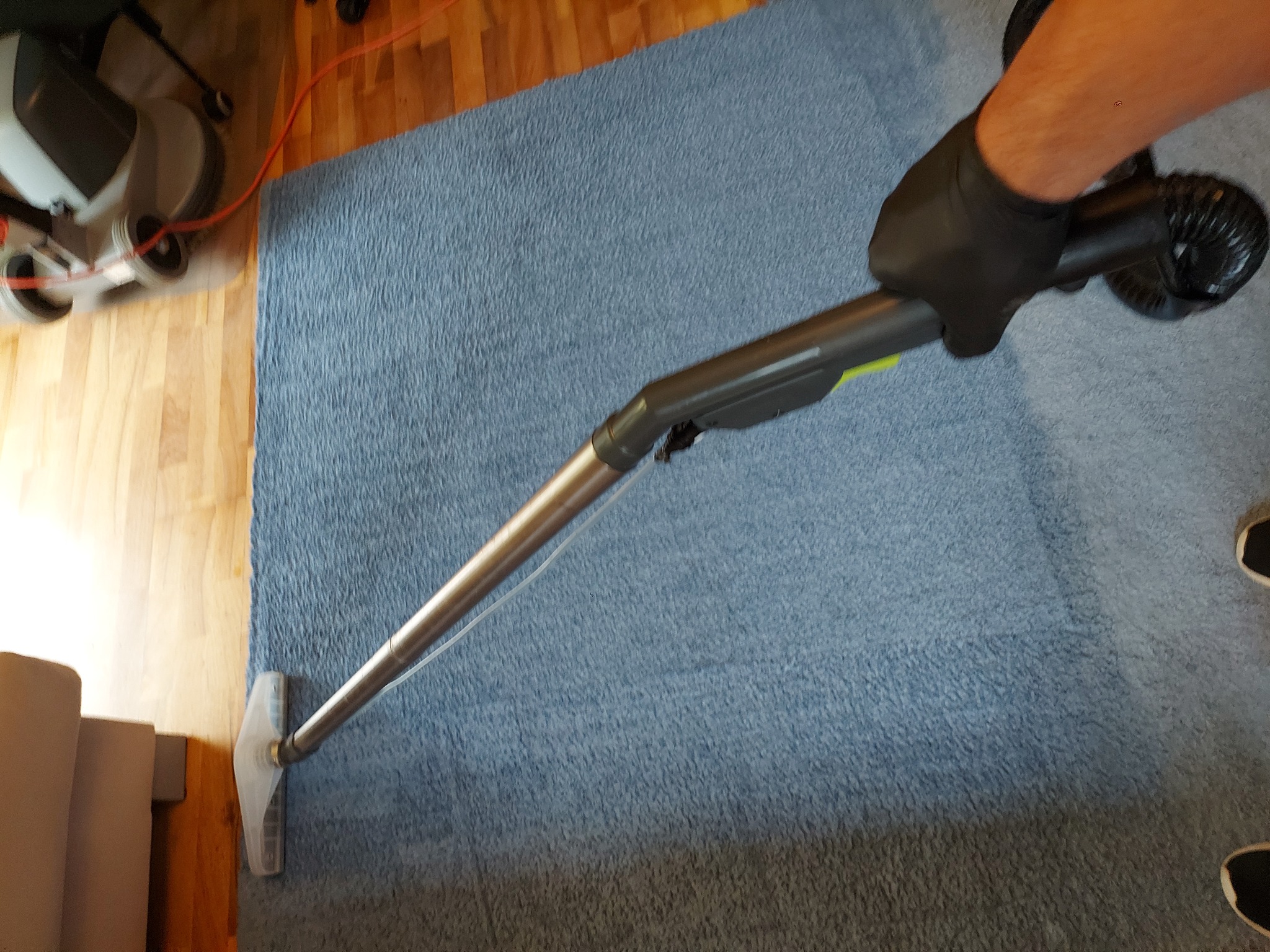 Czyszczenie niebieskiego dywanu za pomocą odkurzacza piorącego z metalową rurą i przezroczystą dyszą, widoczne drewniane panele podłogowe i fragment sofy.