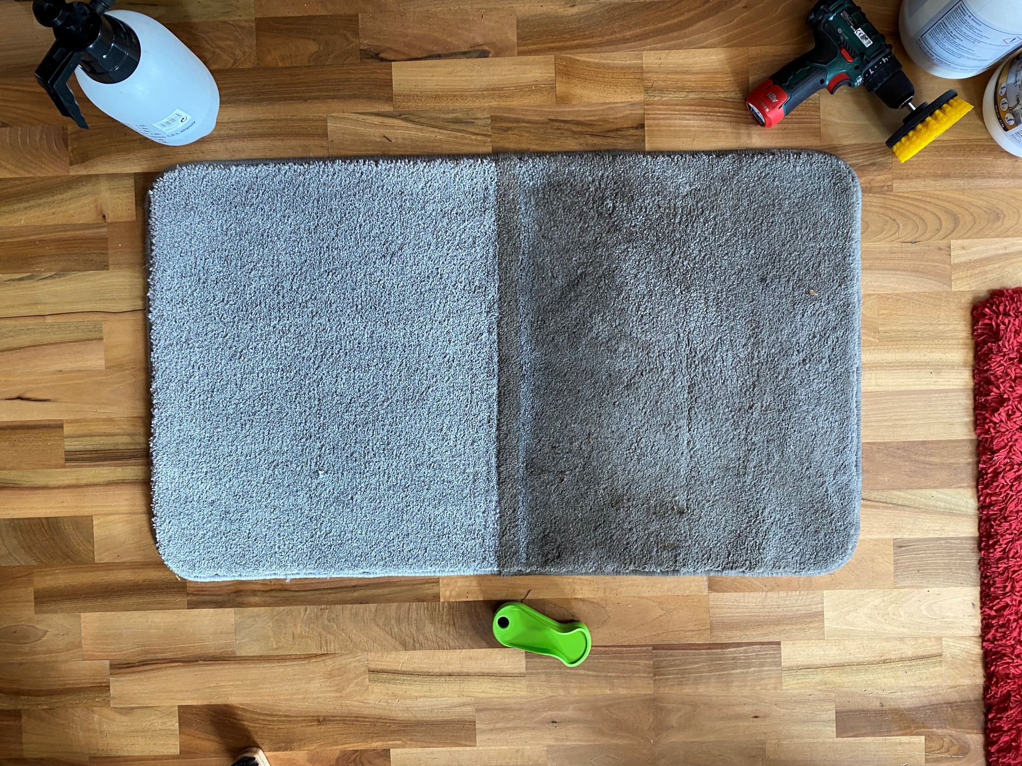 Dywanik z widoczną różnicą czystości: lewa strona jaśniejsza, prawa strona ciemniejsza, obok butelka z rozpylaczem, wiertarka z nakładką czyszczącą i zielony skrobak na drewnianej podłodze.