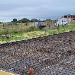 Zbrojenie płyty fundamentowej z wystającymi prętami i rurami kanalizacyjnymi, widok z góry na plac budowy z domami w tle, pochmurne niebo.