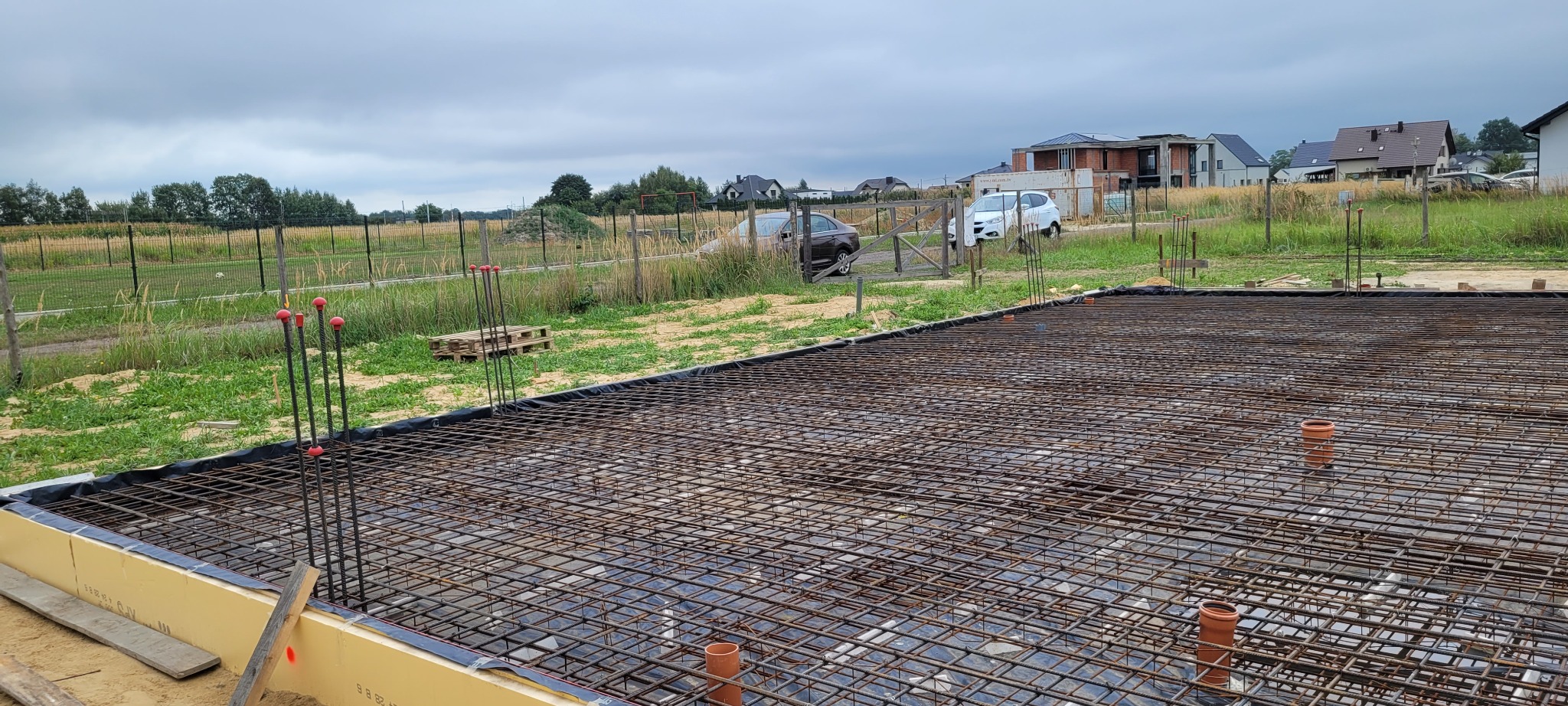 Zbrojenie płyty fundamentowej z wystającymi prętami i rurami kanalizacyjnymi, widok z góry na plac budowy z domami w tle, pochmurne niebo.