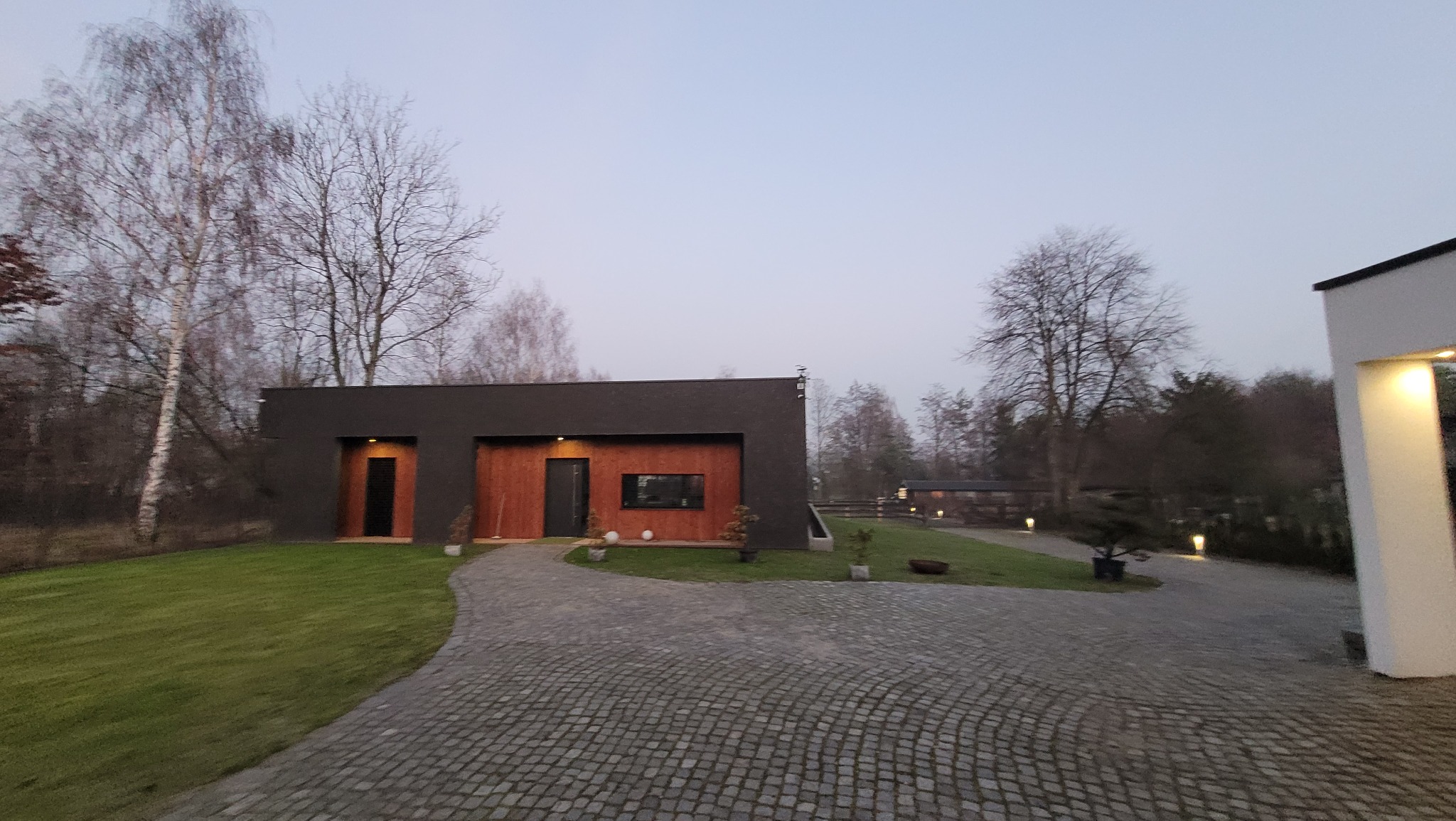 Nowoczesny dom z drewnianą elewacją i brukowanym podjazdem, otoczony zielenią i drzewami. Minimalistyczny design, oświetlenie zewnętrzne. Ujęcie z poziomu gruntu.