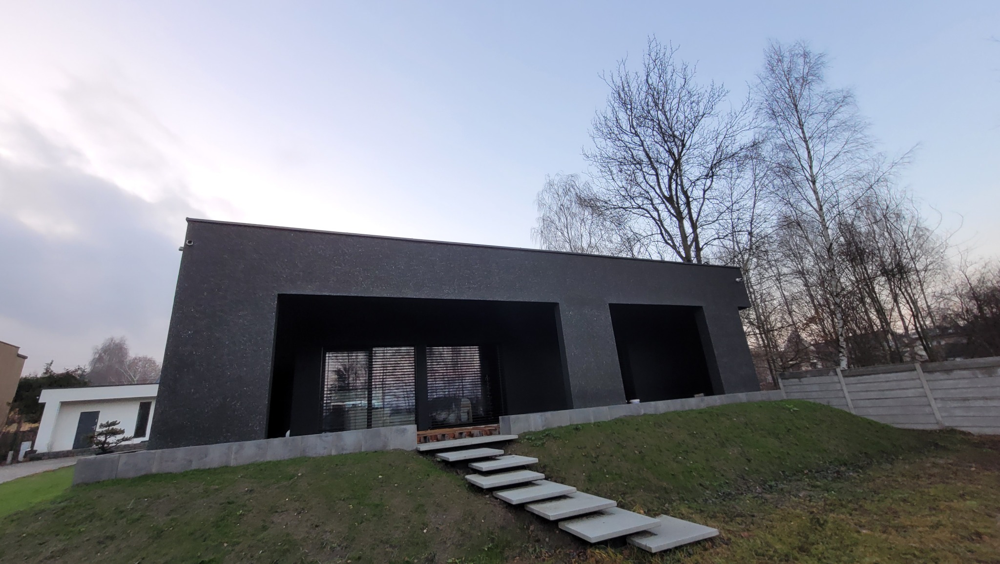 Nowoczesny, czarny dom jednorodzinny z betonowymi schodami prowadzącymi do wejścia. Minimalistyczna architektura, surowe wykończenie elewacji, widok z dołu.