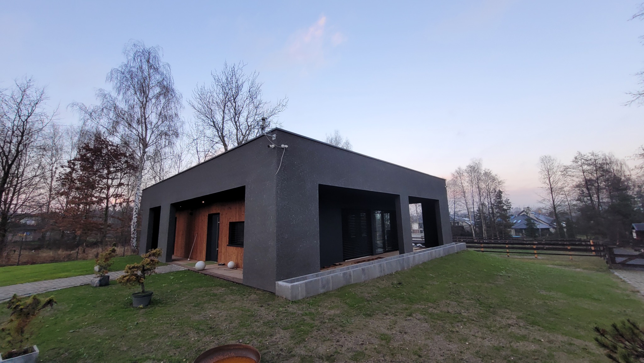 Nowoczesny, parterowy dom z ciemną elewacją i drewnianymi akcentami, otoczony zielenią. Widoczne elementy architektury krajobrazu i system monitoringu.