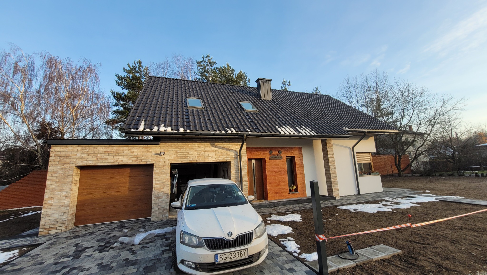 Nowoczesny dom jednorodzinny z garażem, wykończony cegłą i drewnem, z ciemną dachówką i białym samochodem Skoda na podjeździe. Częściowo ośnieżony teren.