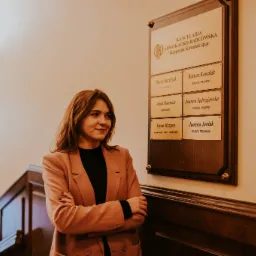 Portret kobiety w różowym żakiecie stojącej na tle tablicy informacyjnej Kancelarii Adwokacko-Radcowskiej z wypisanymi nazwiskami adwokatów i radców prawnych.