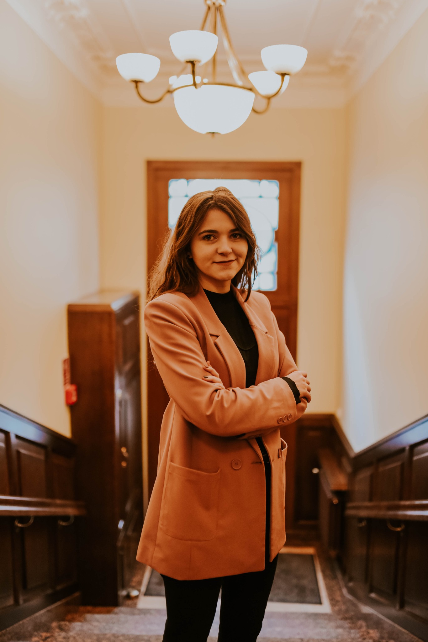 Portret kobiety w brzoskwiniowym żakiecie stojącej na schodach w eleganckim wnętrzu z drewnianą balustradą i stylowym żyrandolem.
