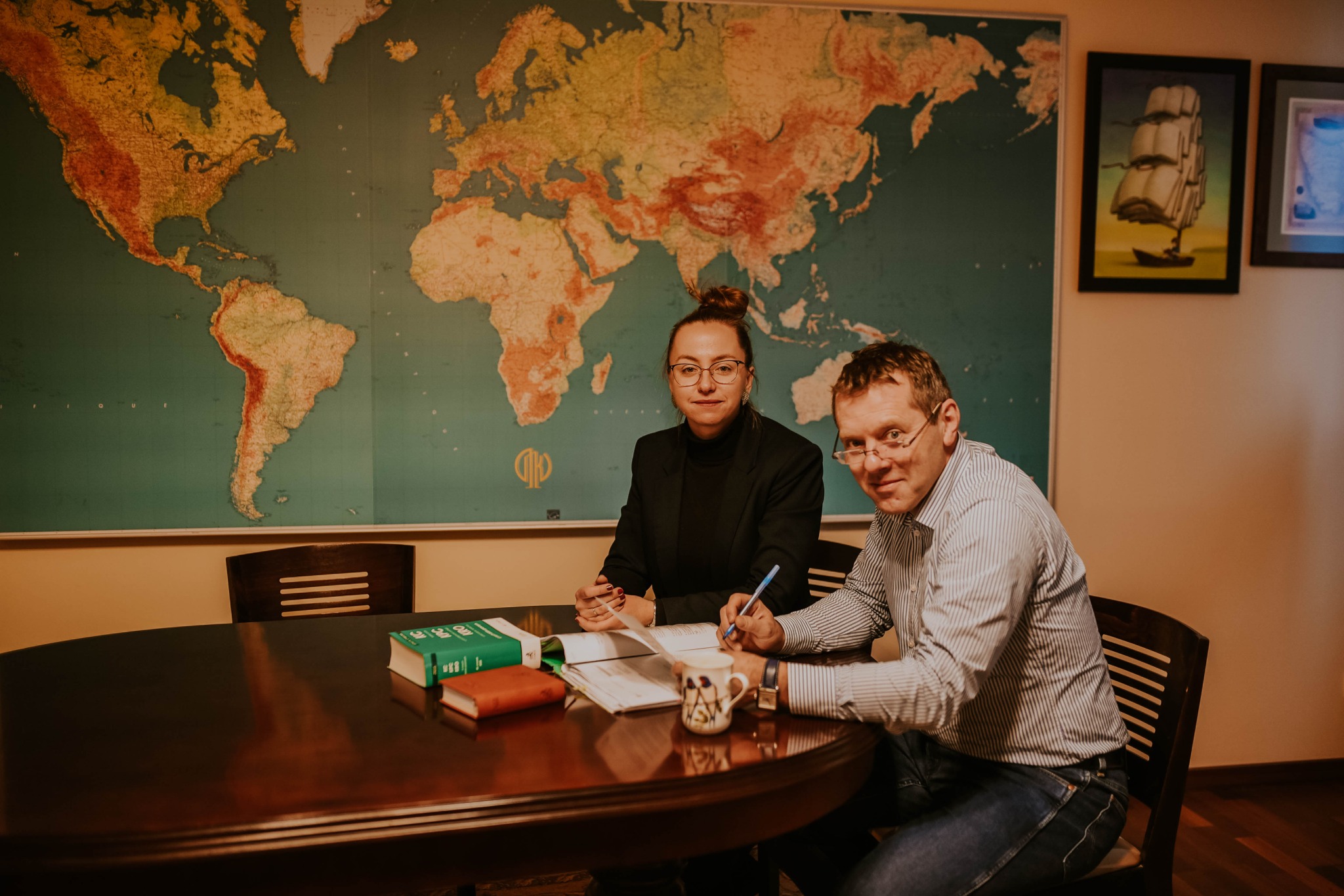 Dwie osoby w okularach przy okrągłym stole z dokumentami, książkami i kubkiem, na ścianie mapa świata i obrazy